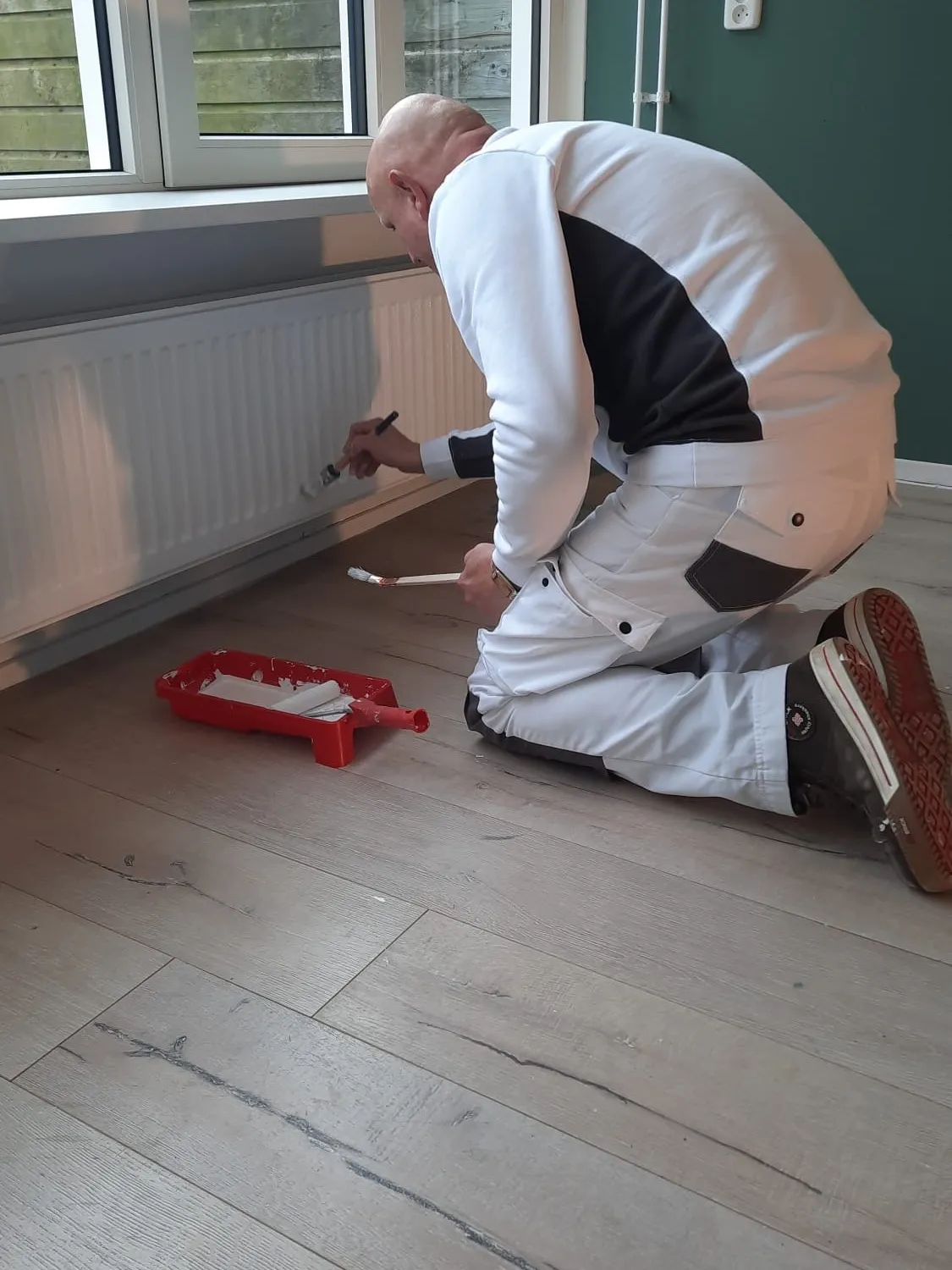 Schilder werkt geknield het houtwerk onder een radiator bij